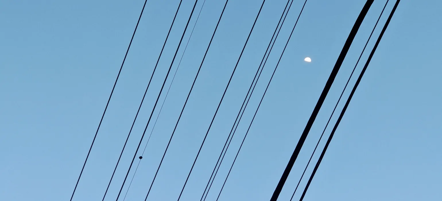 Several power lines of varying sizes appear as black lines covering the center third of the image, rising from the bottom left to the top right across a pure blue sky. In the bottom left, on one line sits a bird, far enough away that it is a small blob. In the top right, centered between two of the lines, is the bright gibbous moon.