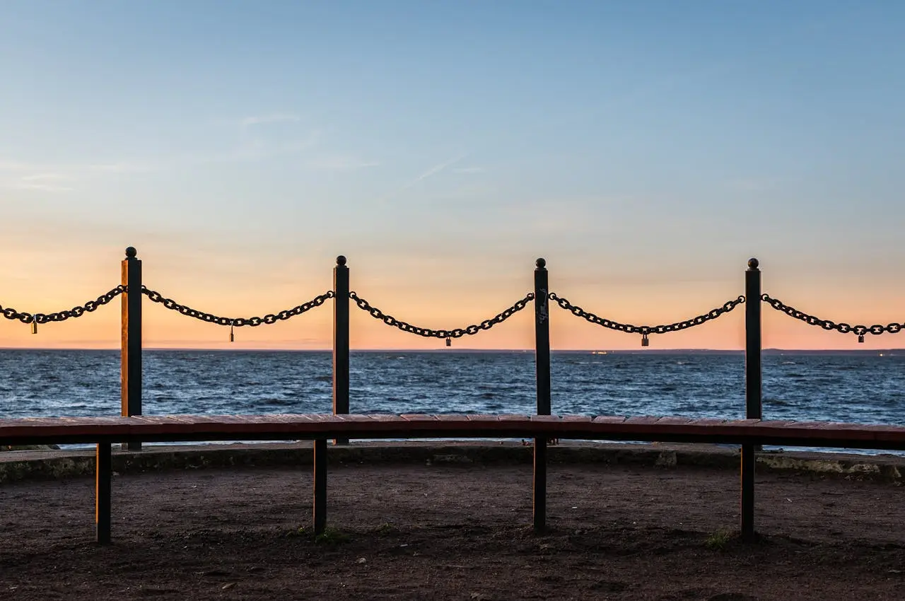Four metal poles connected by chains with padlocks at the center stand against the sea, with a blue-orange sunset above.