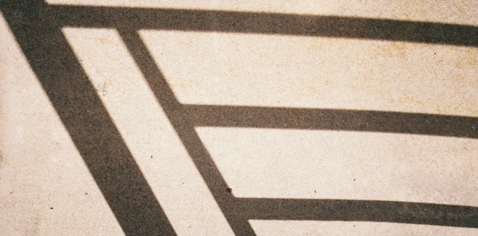 A shadow draws sharp lines on brightly lit concrete. On the left side is a thick line descending across the full height diagonally, connected at the top to a subtly curved line which spans the full width of the image. It is connected by a thinner parallel diagonal line, which has two more horizontal lines equally spaced across the image.