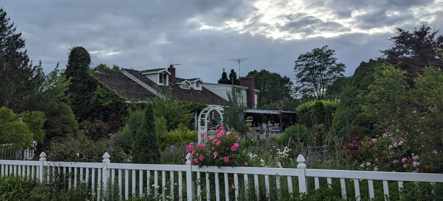 The bottom of the image is spanned by a white fence, behind which a lush green garden and house sit. Above is a moody, cloudy sky. There are prominent pink flowers in the center, while there are white and dark purple flowers sprinkled through the rest of the garden.