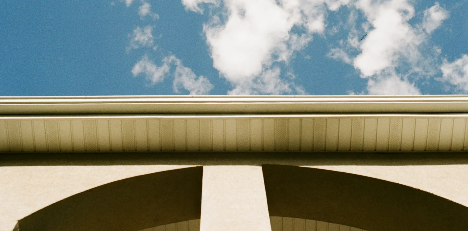 A light blue cloudy sky is broken horizontally down the center of the image by the bottom side of a light tan roof viewed from below at a high angle. Below the roof, under a thin shadow from the roof, a thick stucco wall has a center beam and curves down away from it into two arches.