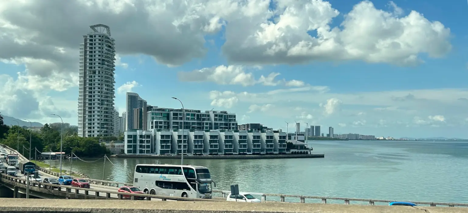 Cars drive across a road bridge over light blue water. Above are several large hotels, and white clouds against a light blue sky.