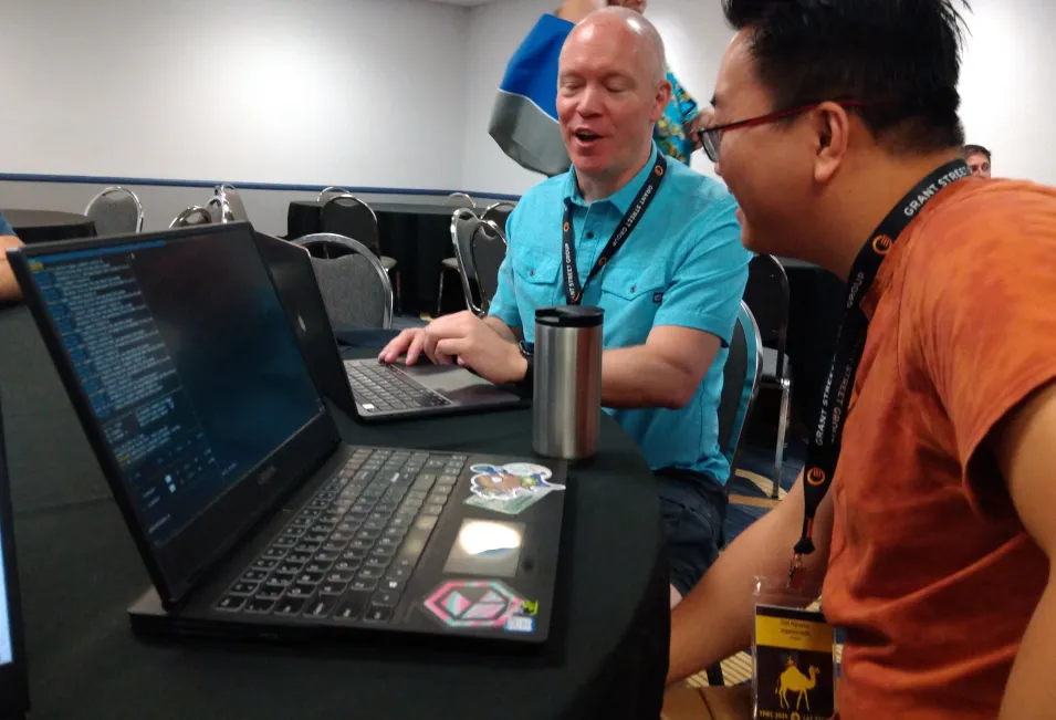 Two men sit at a table in front of their computers, one shows the other something and has a slight smile