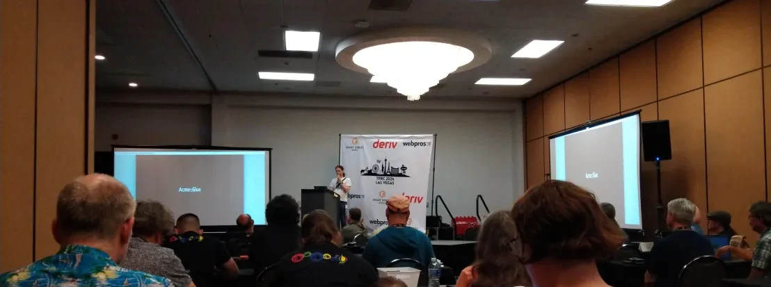 A conference room with around 30 people visible watching a speaker talk, in front of a TPRC banner.