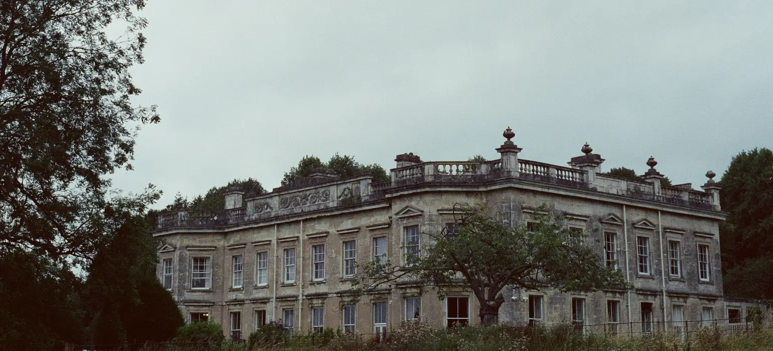 A corner angle of a 100-200-year-old house sitting among green trees against a dark blue sky. Its many windows are neatly arrayed, and well maintained.