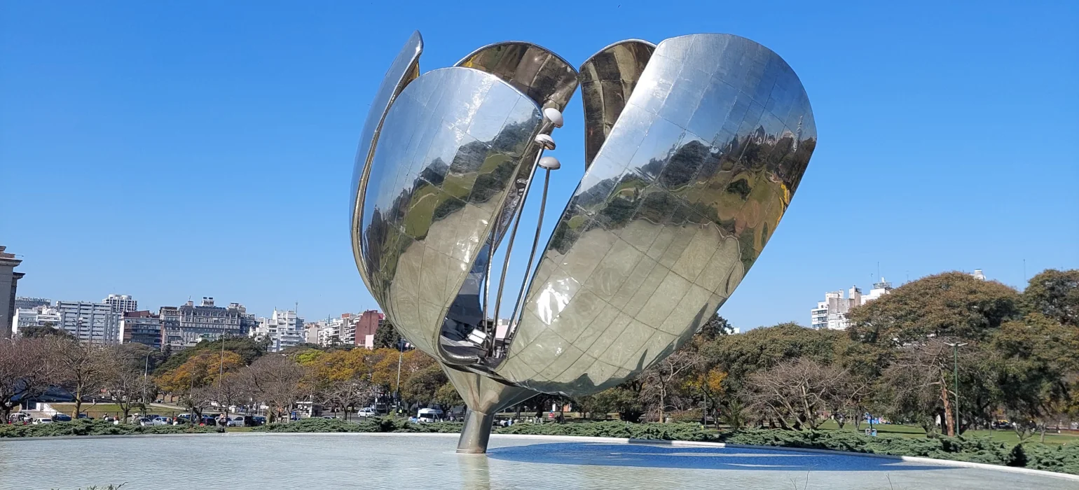 A metallic, reflective, flower-shaped sculpture in a shallow pool of water. The pool is surrounded by a park, with a city skyline in the background.