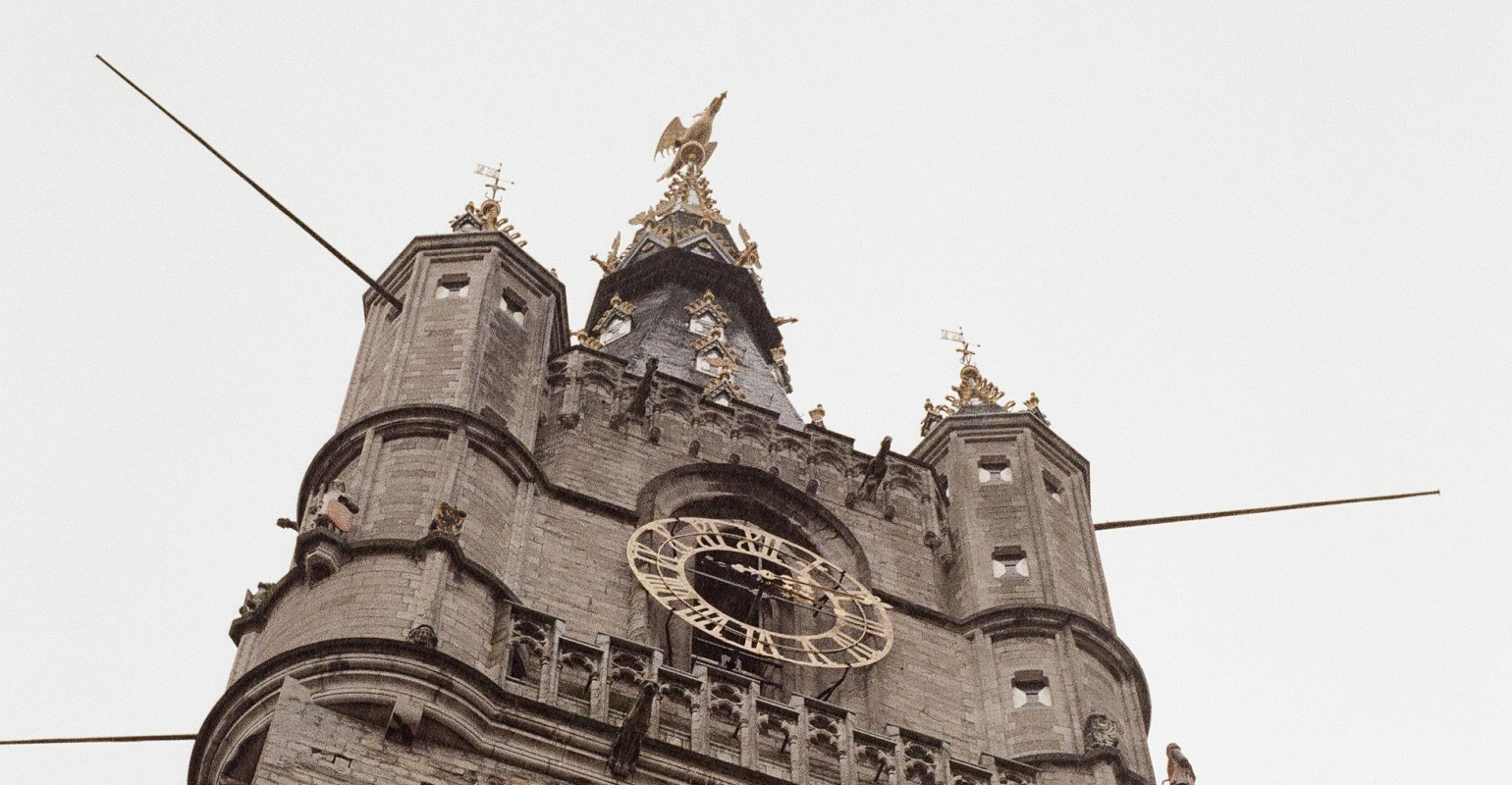 A low-angle view of an old European church clock tower. The square tower with rounded corners is ornamented with gothic styling, and topped with a golden eagle.