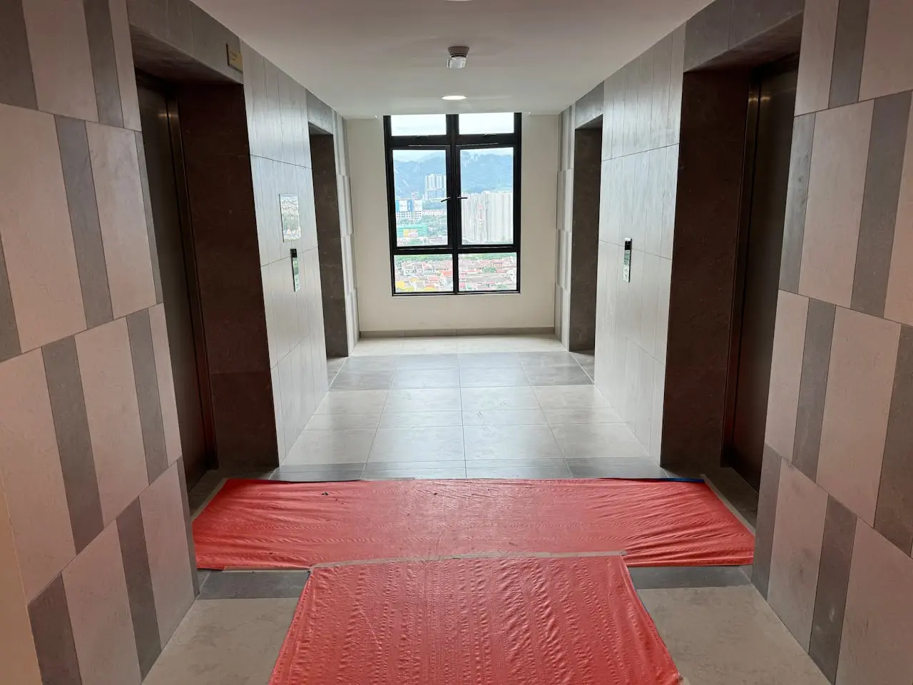 Two elevators on either side of a hall, with orange construction plastic stretched out in front of the entrances. At the end of a hall is a window looking out over a city.