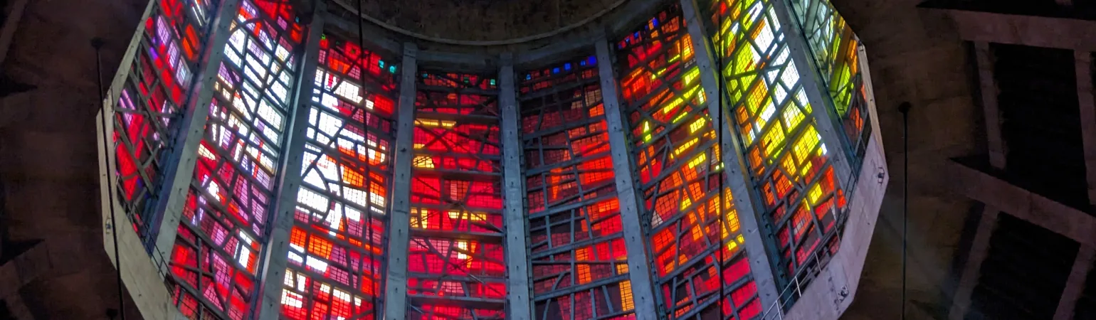 A low angle view of a gridded ceiling, leading to a large stained glass cylindrical top, which is lined with multicolored stained glass in a highly varied arrangement.