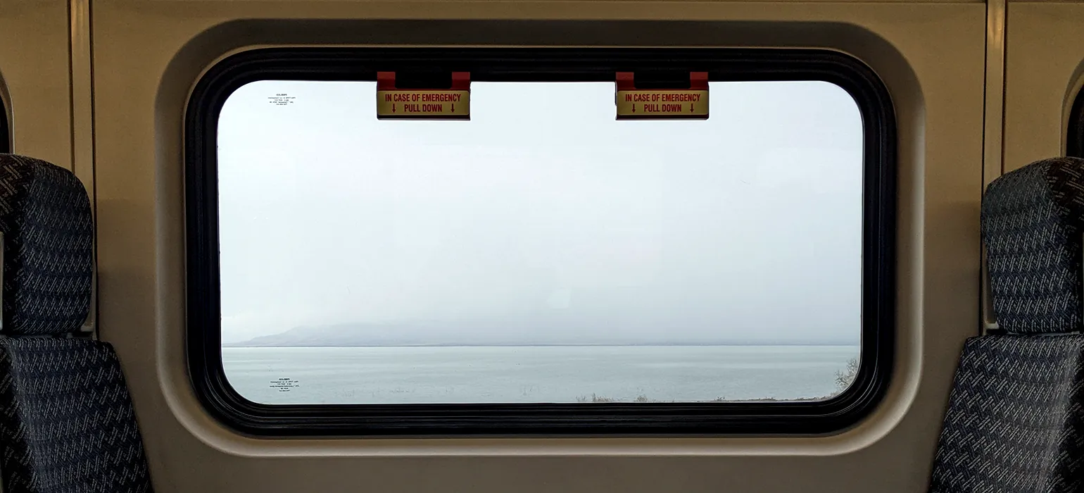 A misty lake with mountains hidden by fog and clouds is viewed through a rectangular train window, straight on.