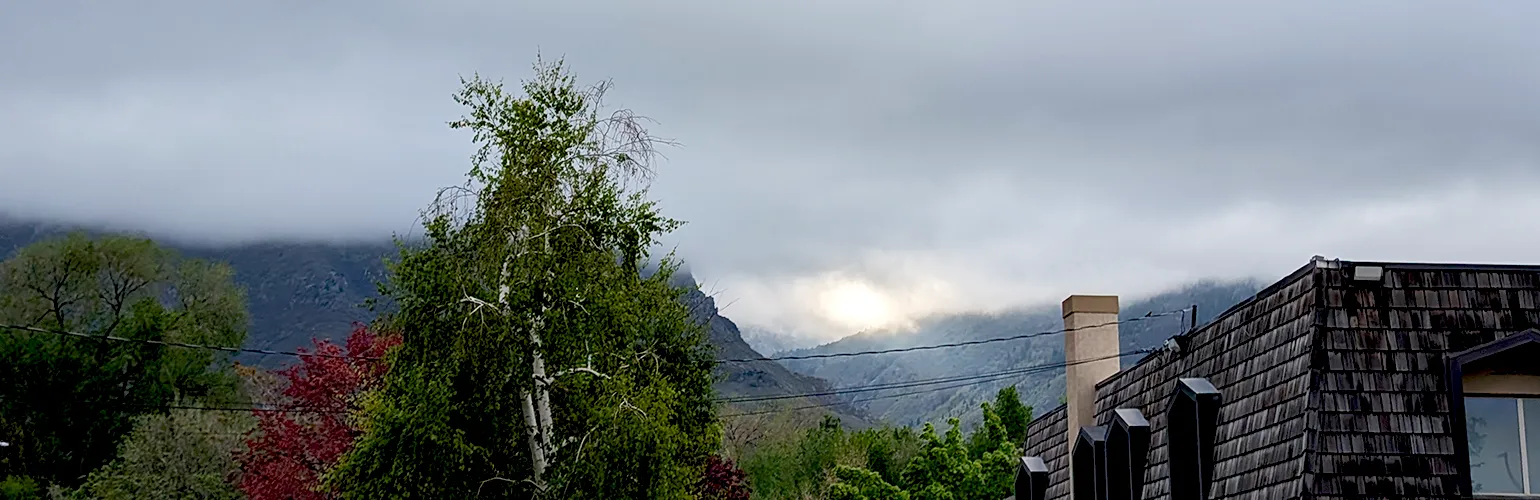 The camera looks over houses and green and red trees at a mountains just visible below a thick layer of clouds. In the center of the image is a canyon lit by sunlight, which peeks through the clouds above the canyon.