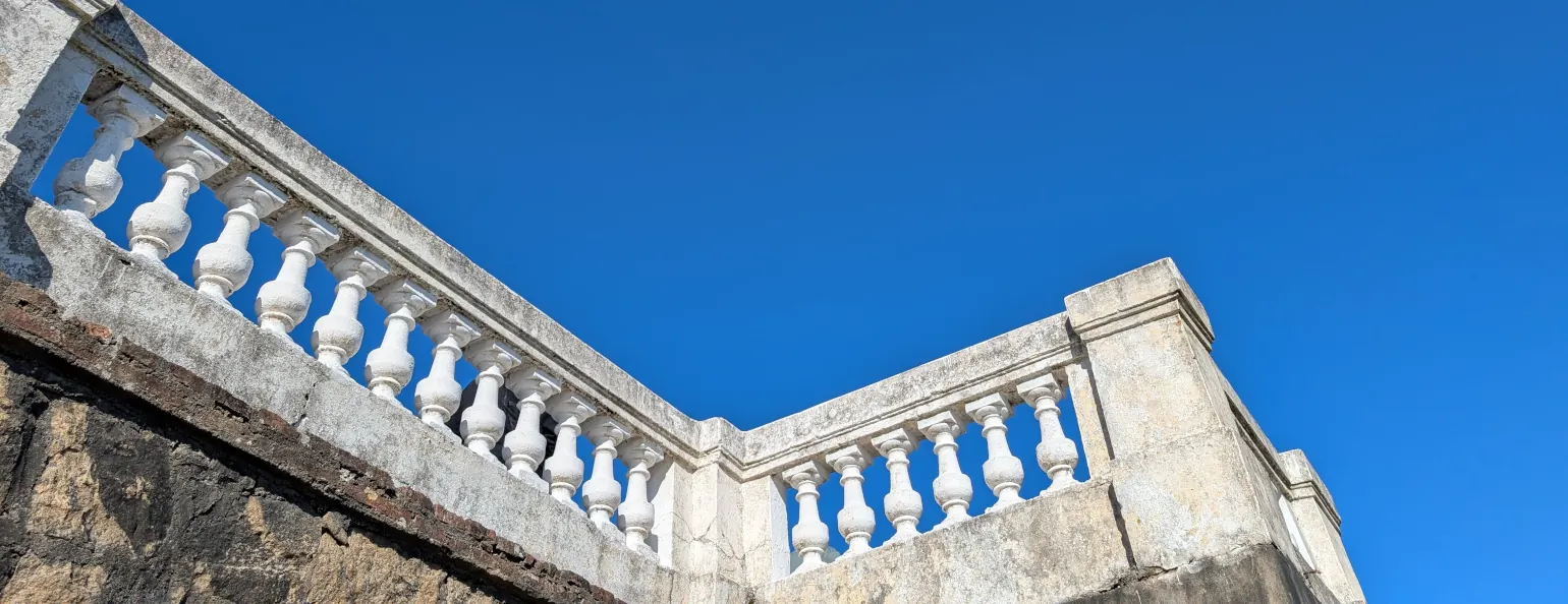A white plastered railing from a low angle, behind which is a pure blue sky. The railing comes down and to the right from the top left corner of the image, then has a right angle in the center of the image, coming back up and to the right for another quarter of the image before stopping.