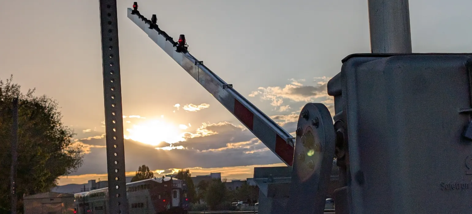 A rail barrier is in the process of opening, creating a strong line from the bottom right to the top left of the image. Behind is a train on the tracks in from of a deep gold sunrise