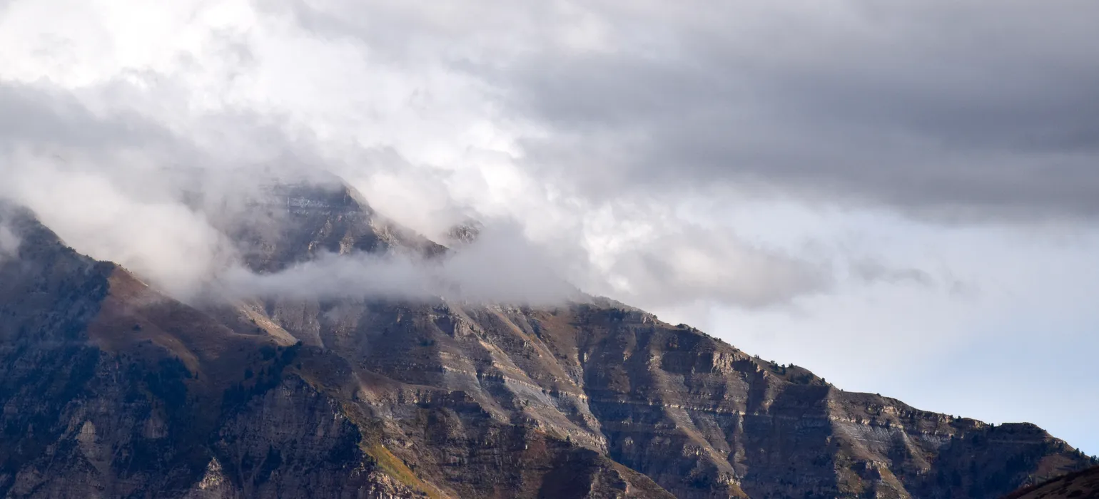 A tall peak coated in brown and green short shrubbery, broken up only by sharp faces of rocky cliffs, sits behind wispy clouds, against an overcast backdrop. The ridgeline descends from the top left downward to the right, splitting the image in half