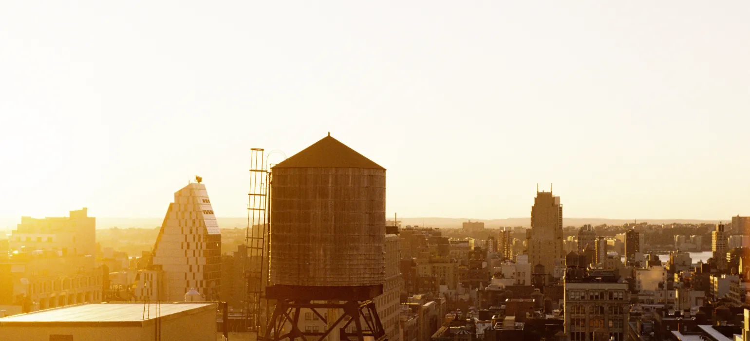 An orange sun sets over New York City, viewed from the top of a building in Manhattan. A water tower is visible in front of buildings which reflect bright light on the left side, with shadows on the right side.