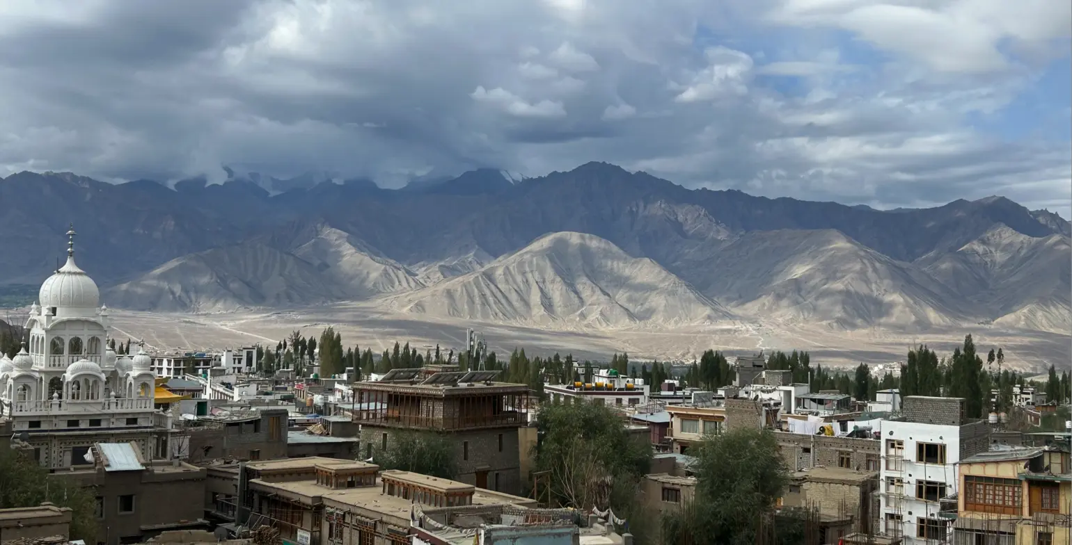 Apartment buildings and a domed white Indian building in Leh city, India sit among trees in front of a layered mountain range, capped with clouds.