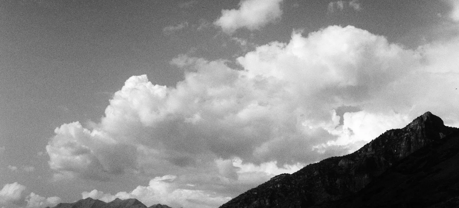 A black-and-white image of clouds hanging over a sharp mountain, jutting from the bottom right of the image, and a longer mountain ridge further back in the bottom left of the image.