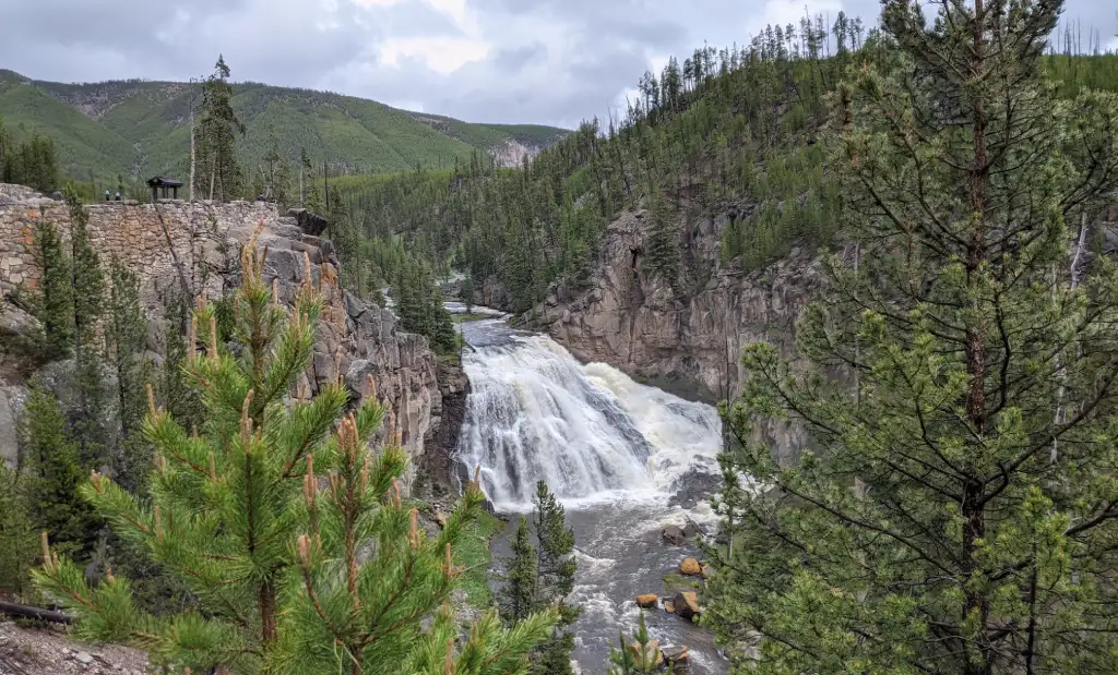A wide waterfall cascades through a rocky, forested canyon surrounded by pine-covered hills under a cloudy sky.