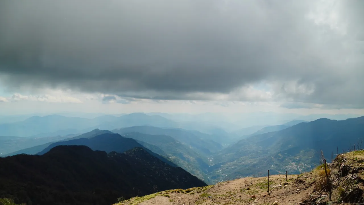 A panoramic view from a mountain overlooks a vast valley stretching to the horizon under a stormy sky.