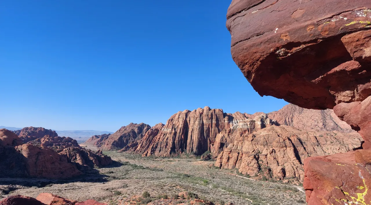 A sweeping desert canyon stretches, framed by towering red sandstone formations and sparse desert scrub under a cloudless blue sky.
